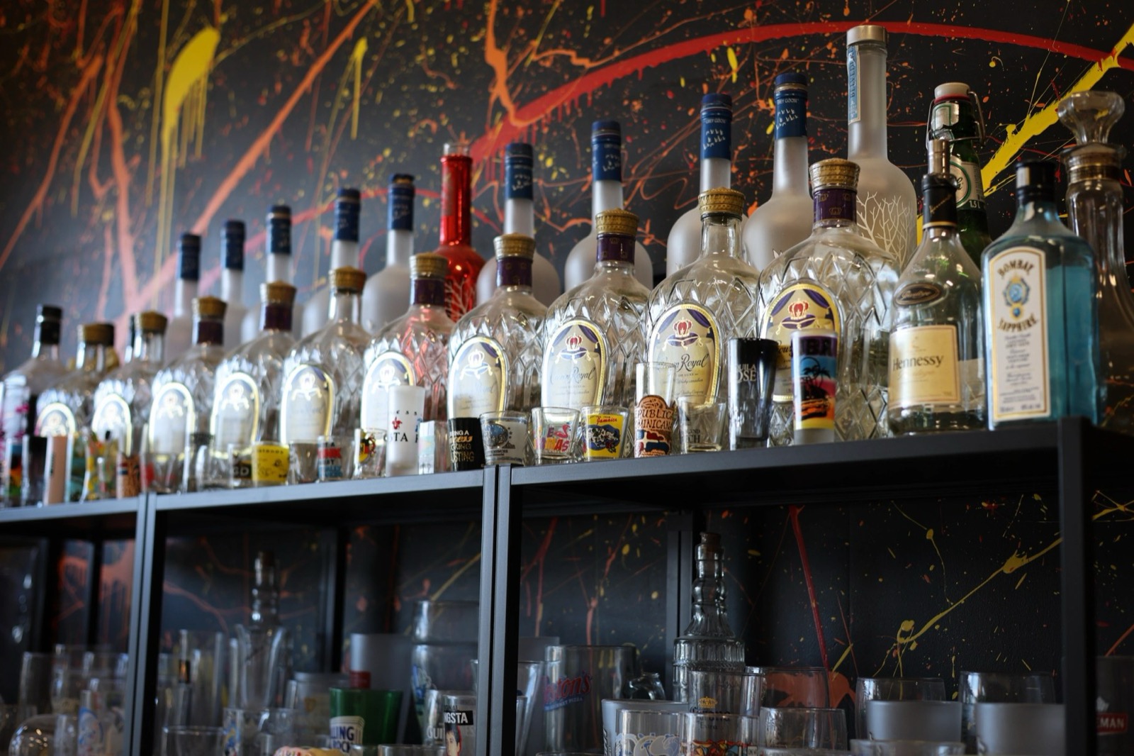 Shelves of glass bottles and shot glasses against a wall covered in flung red, yellow and orange paint