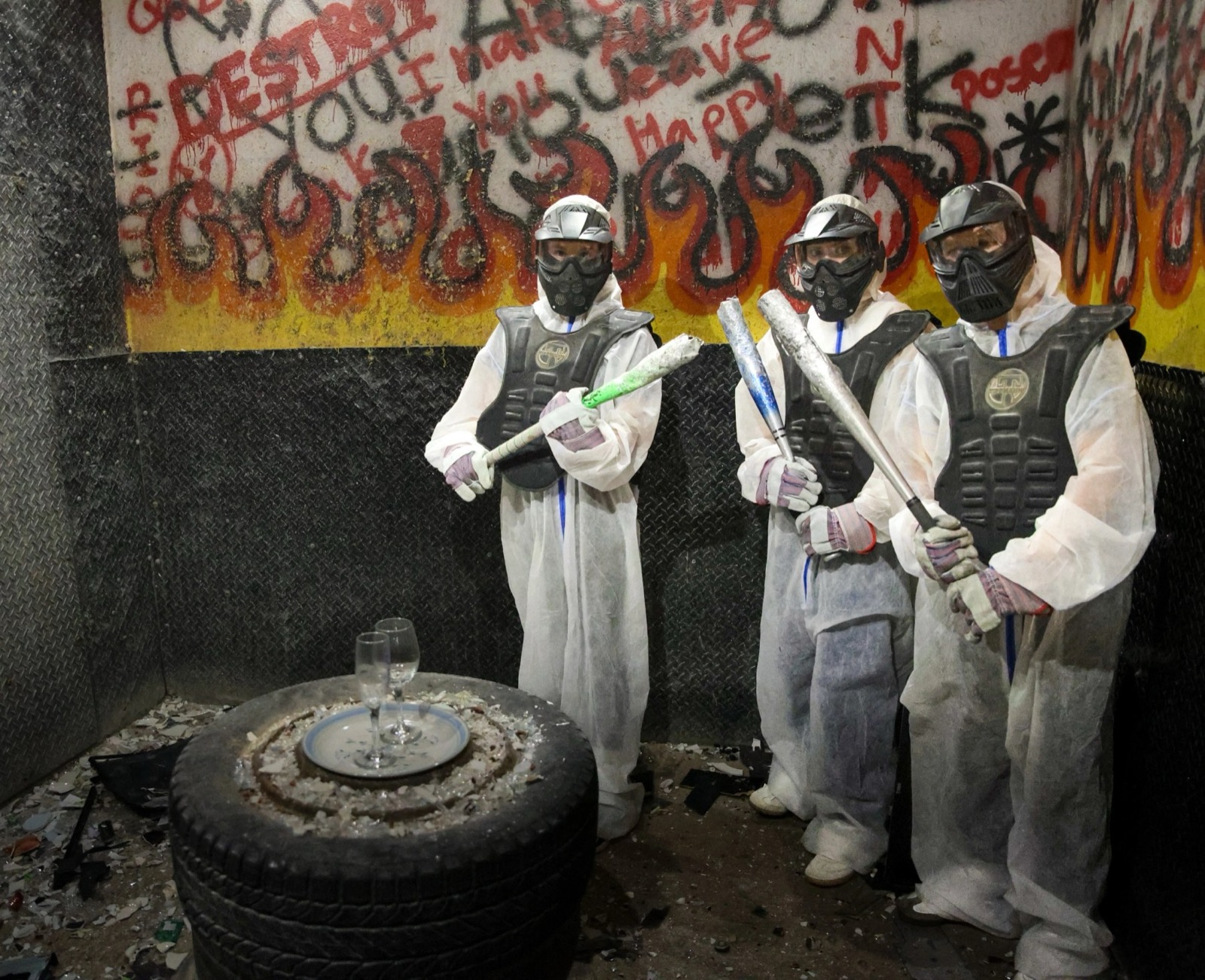 Three people in full safety gear holding bats in front of a graffiti wall painted with flames