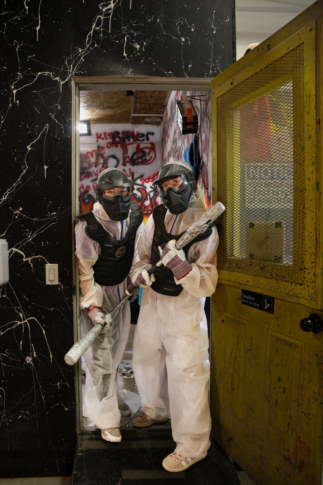 Two people in white coveralls, chest protectors and face masks standing in the rage room doorway holding bats